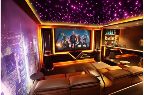 Home theatre room featuring a Starefx ceiling with purple starlights, cove lighting, a large movie screen, lounges, a bar, and a wall fitted with a backlit image of the NYC skyline.